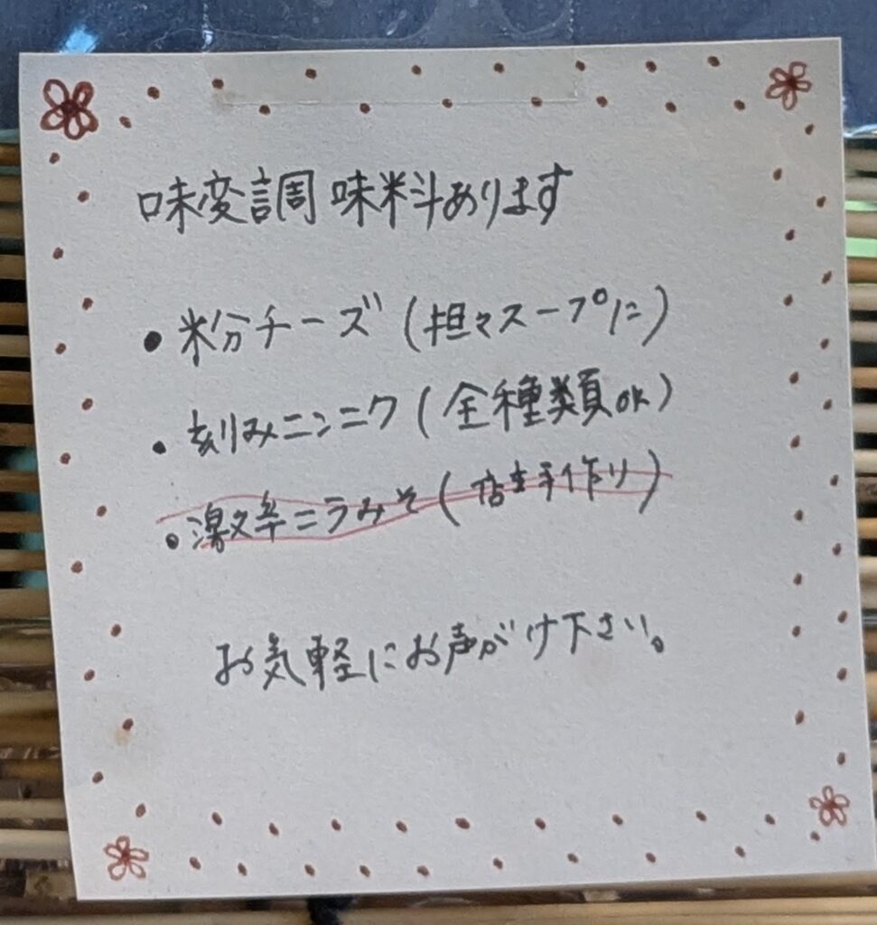『ラーメン 大器』の店内に掲示された味変調味料の案内POP。粉チーズや刻みニンニクの提供を知らせる手書きメモ。